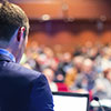 A speaker lectures to a crowded conference room.